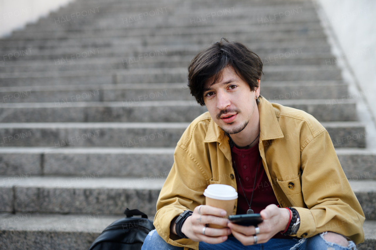 A portrait of young man sitting on staircase outdoors in city, using smartphone