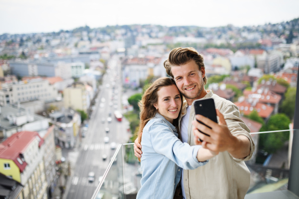 A happy young couple in love standing outdoors on balcony at home, taking selfie with smartphone.