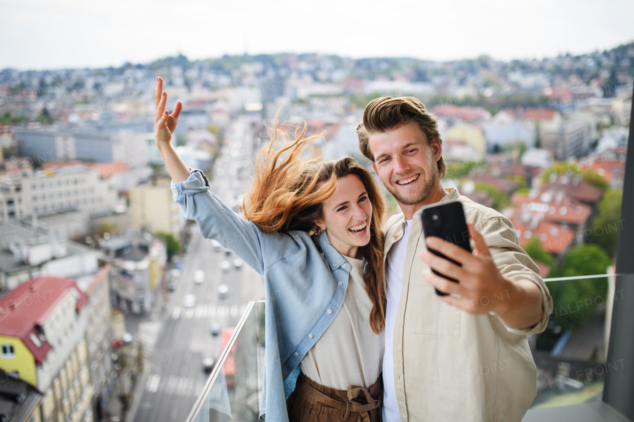 A happy young couple in love standing outdoors on balcony at home, taking selfie with smartphone.