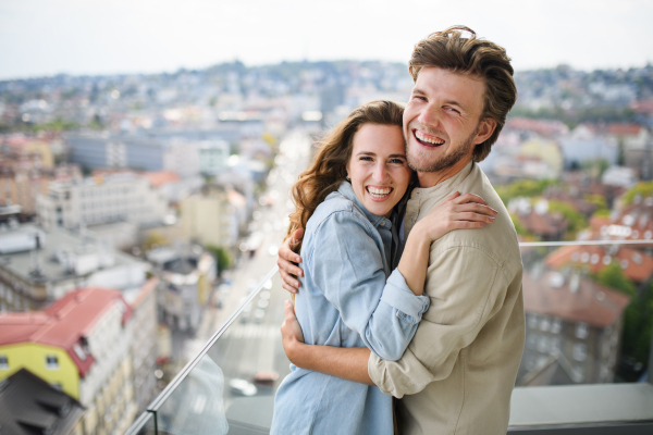 A happy young couple in love looking at camera outdoors on balcony at home, hugging.