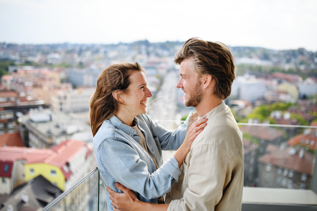 A happy young couple in love standing outdoors on balcony at home, looking at each other.