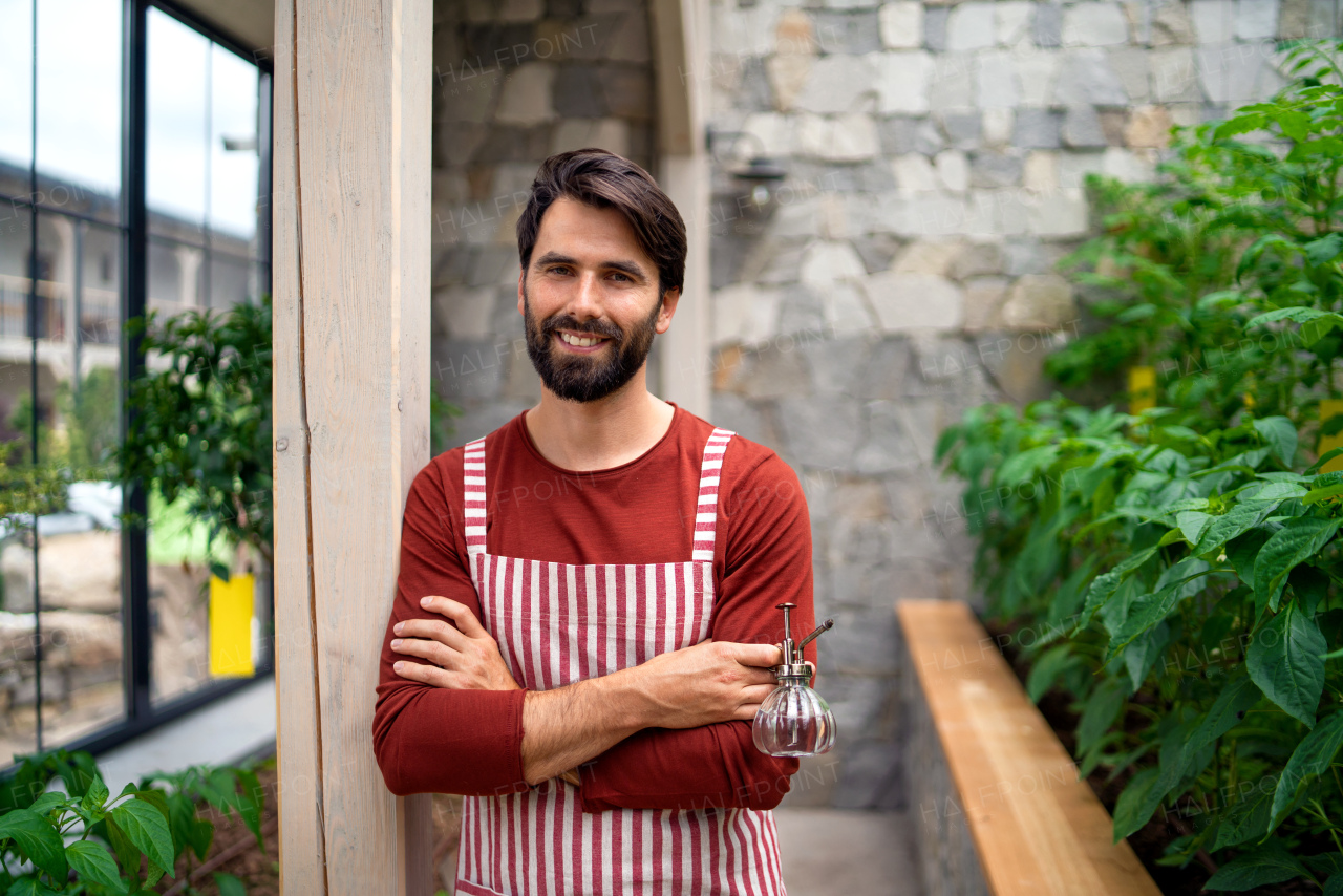 Portrait of man gardener with apron standing in greenhouse, looking at camera.