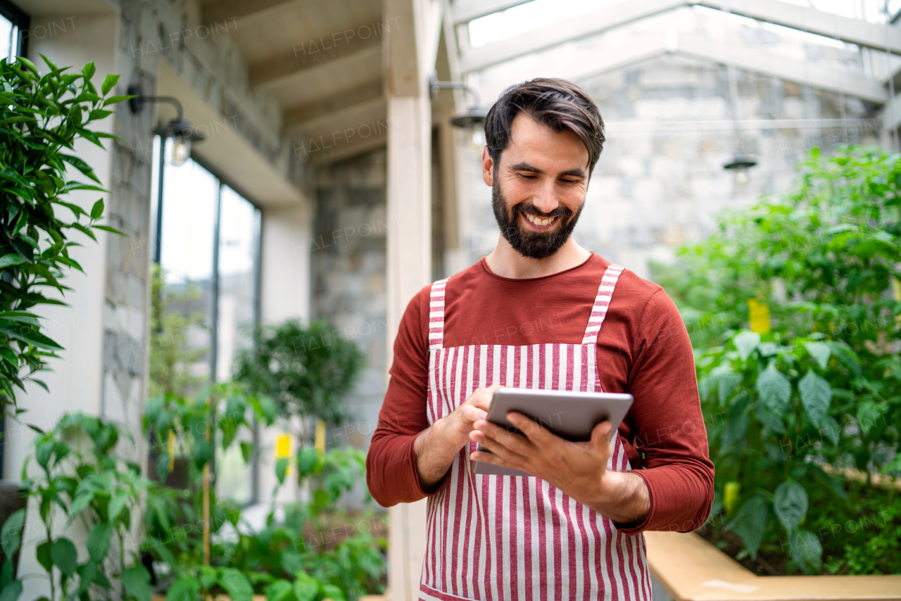 Front view of man gardener with tablet standing in greenhouse, working.