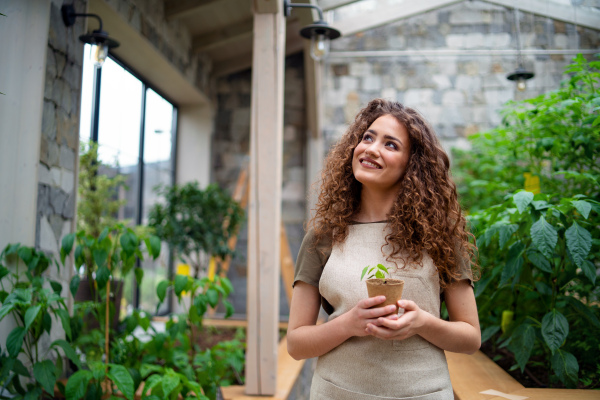 Portrait of woman gardener standing in greenhouse, holding small plant.