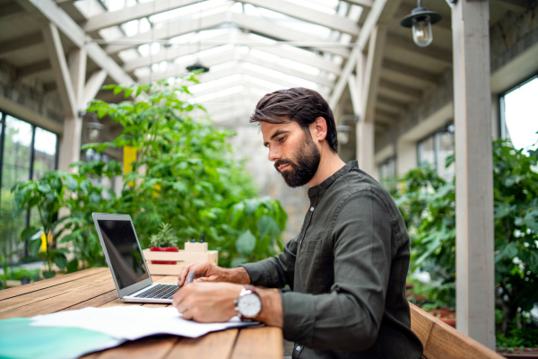 Portrait of young man with laptop sitting indoors in green office, working.