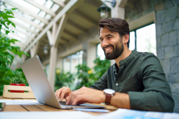 Portrait of young man with laptop sitting indoors in green office, working.