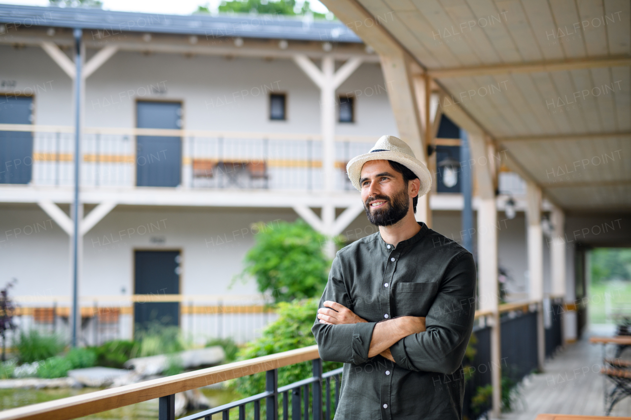 Front view of young man standing outdoors by hotel on holiday, arms crossed.