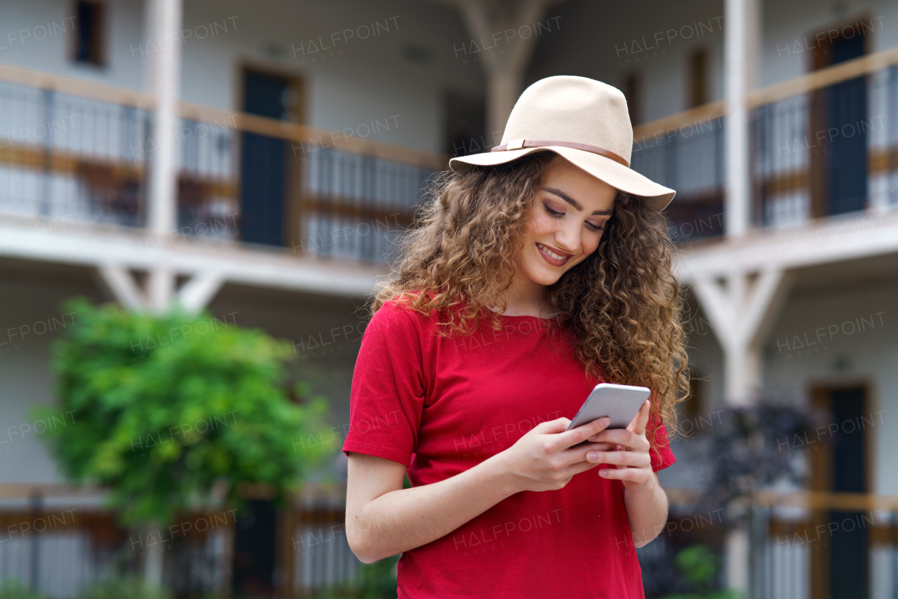 Happy young woman standing outdoors by hotel on holiday, using smartphone.