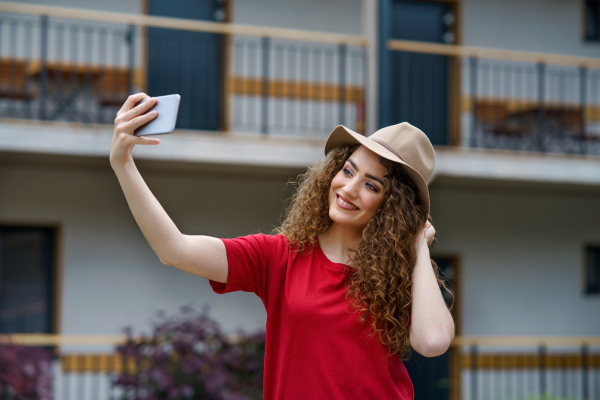 Happy young woman standing outdoors by hotel on holiday, taking selfie.