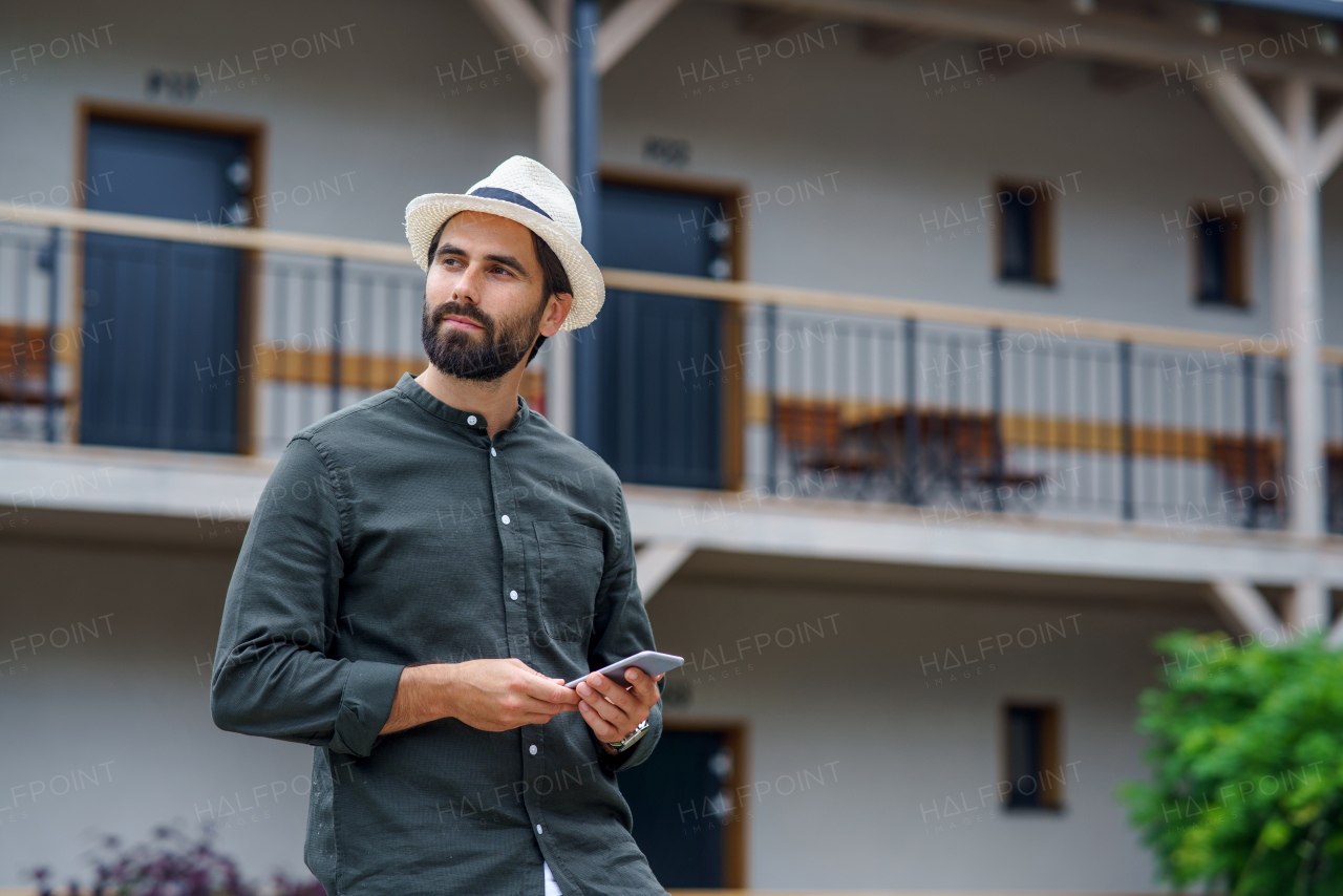 Front view of young man standing outdoors by hotel on holiday, holding smartphone.