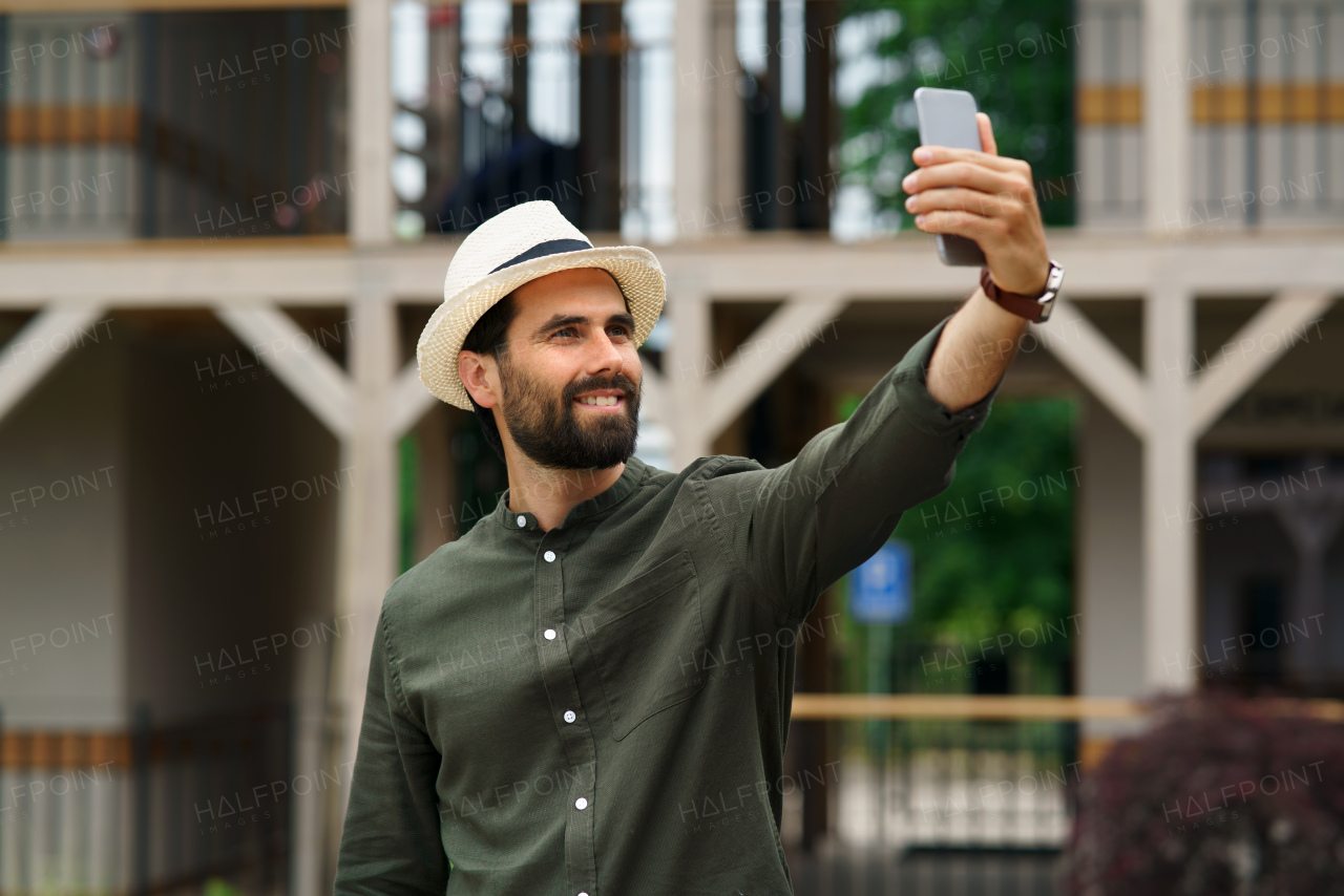 Young man standing outdoors by hotel on holiday, taking selfie by smartphone.