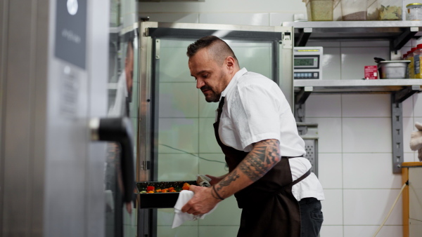 A man chef taking out metal baking sheet with chopped vegetables from oven in restaurant kitchen.