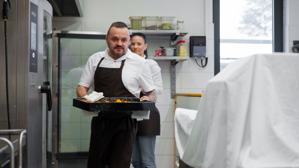 A man chef taking out metal baking sheet with chopped vegetables from oven in restaurant kitchen.