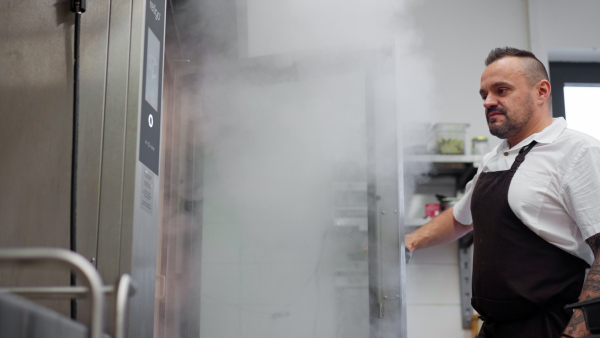 A man chef opening oven and the steam comes out in commercial kitchen.