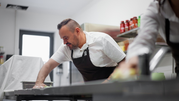 A chef and cook cleaning the workspace after doing dishes indoors in restaurant kitchen.