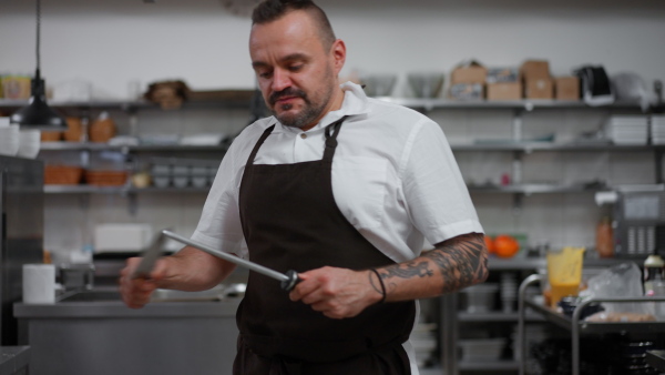 A professional chef sharpening knife and looking at camera in restaurant kitchen.