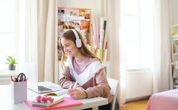 Young happy college female student sitting at the table at home, using laptop and headphones when studying.