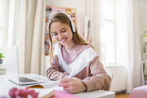 Young happy college female student sitting at the table at home, using laptop and headphones when studying.