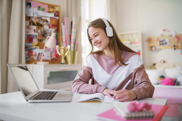 Young happy college female student sitting at the table at home, using laptop and headphones when studying.