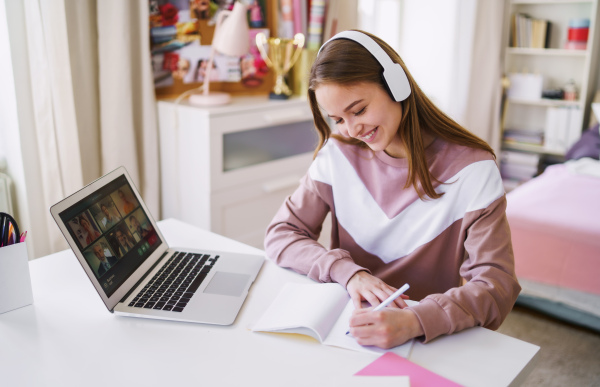 Young happy college female student sitting at the table at home, using laptop and headphones when studying.