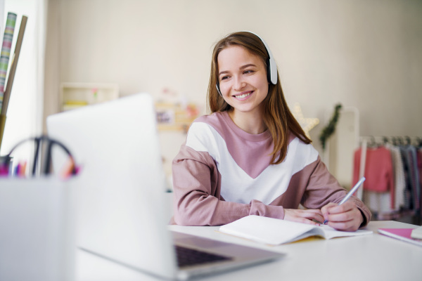 Young happy college female student sitting at the table at home, using laptop and headphones when studying.