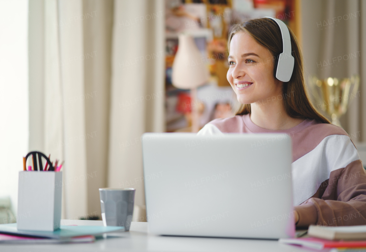 Young happy college female student sitting at the table at home, using laptop and headphones when studying.