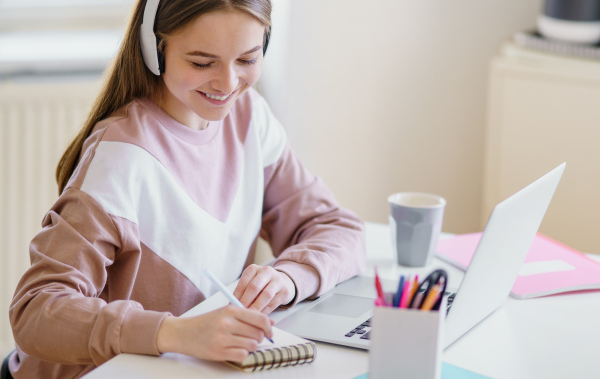 Young happy college female student sitting at the table at home, using laptop and headphones when studying.