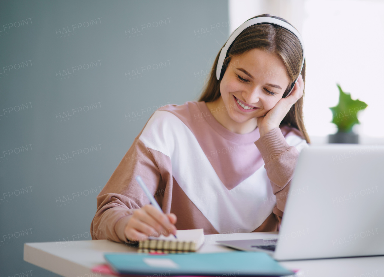 Young happy college female student sitting at the table at home, using laptop and headphones when studying.