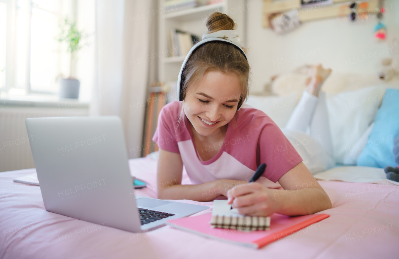 Young female student with face mask and laptop lying on bed, online lesson concept.