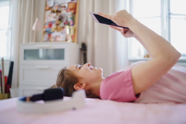 Side view of young girl with tablet on bed, relaxing during quarantine. Copy space.