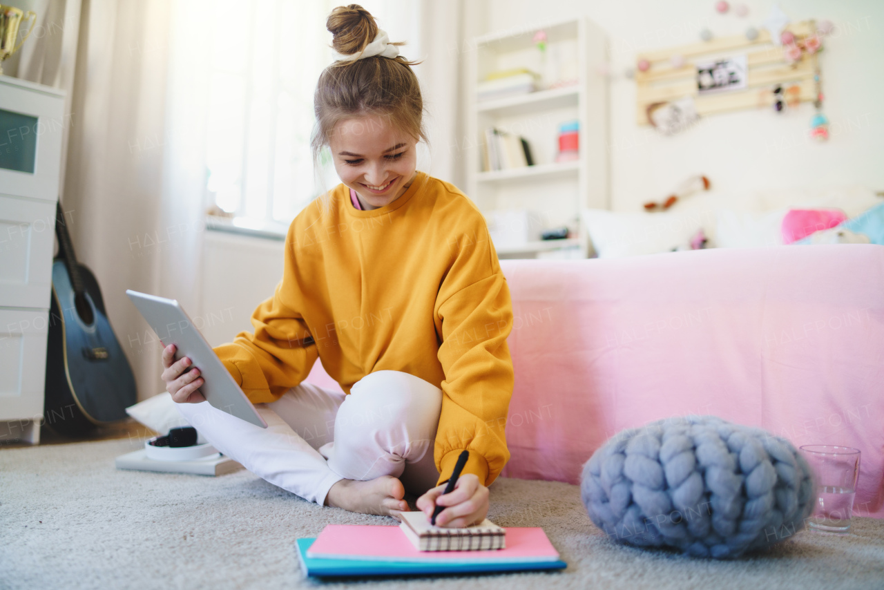 Young girl with tablet sitting and writing on floor, studying during quarantine.