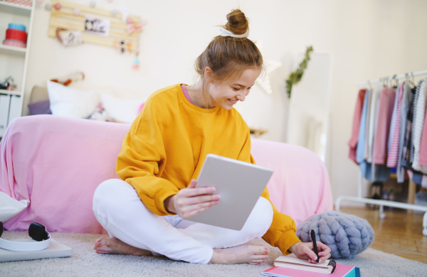 Young girl with tablet sitting and writing on floor, studying during quarantine.