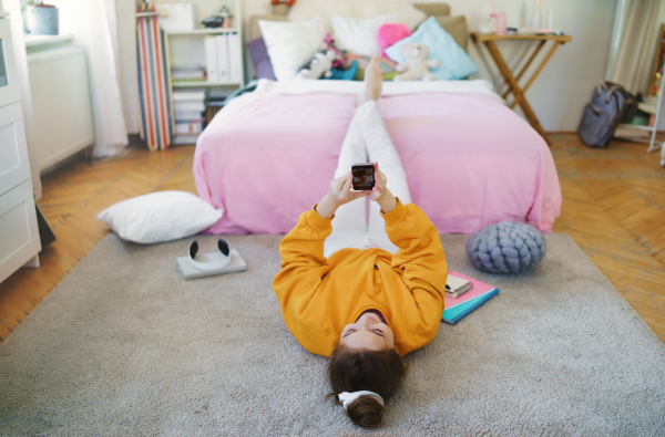 Happy young girl with smartphone lying on floor at home, making video call.