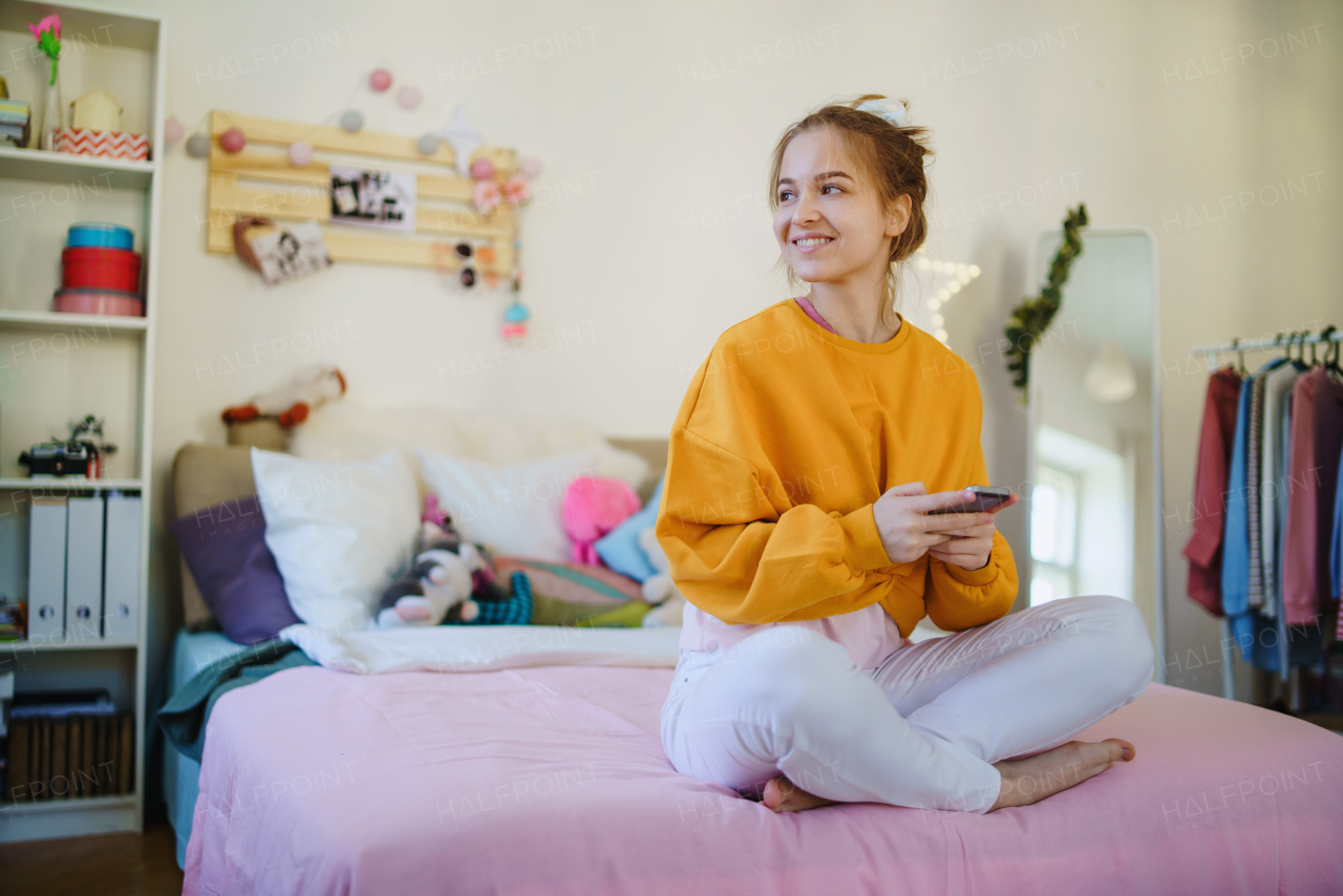 Happy young female student sitting on bed, using smartphone. Copy space.