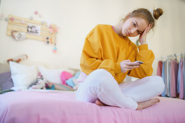 Sad young female student sitting on bed, using smartphone. Copy space.