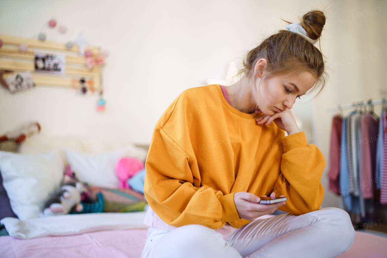 Sad young female student sitting on bed, using smartphone. Copy space.