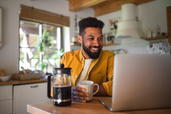 A young man with laptop and coffee working indoors, home office concept.