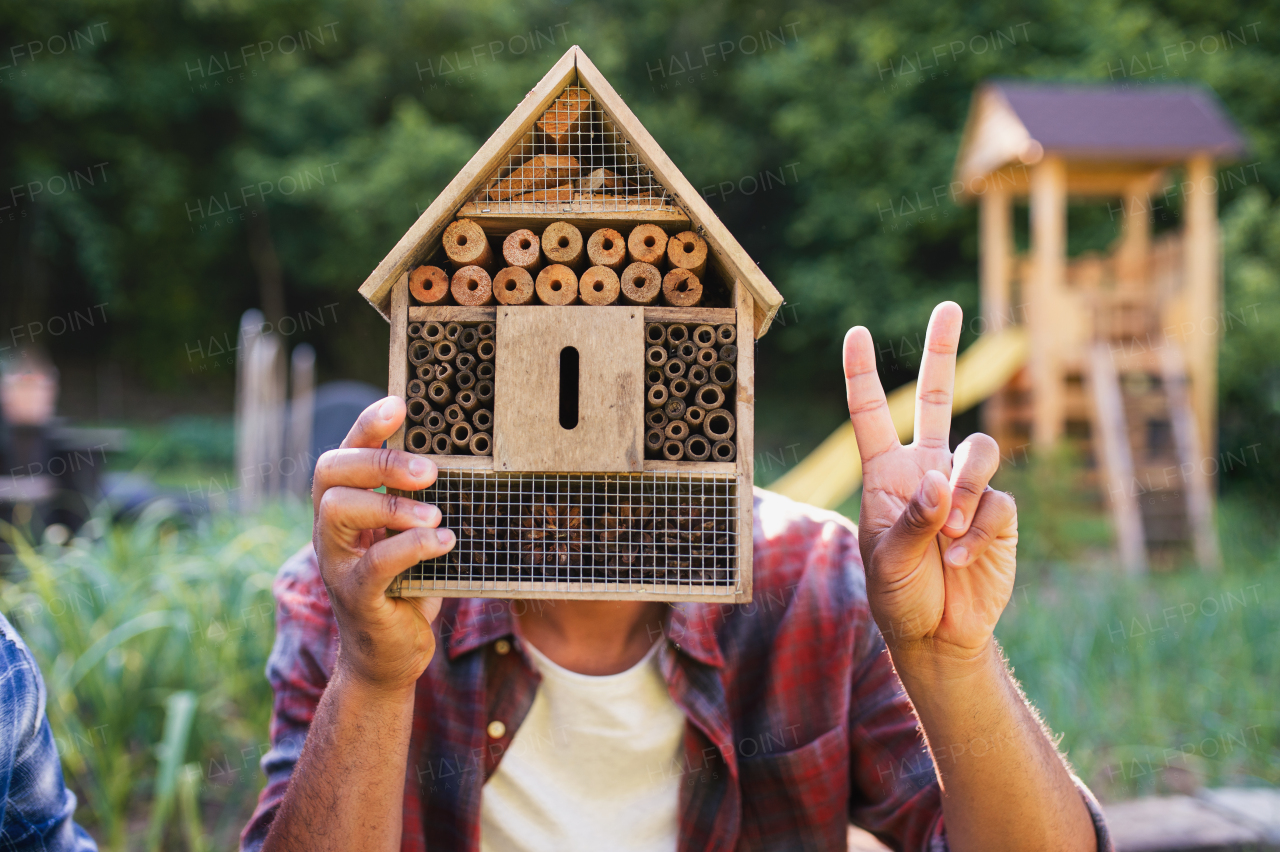 Front view of happy young man with bug hotel spending time outdoors in backyard, showing v sign with fingers.