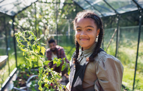 A happy small girl gardening in greenhouse outdoors in backyard, looking at camera.