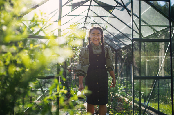 A happy small girl gardening in greenhouse outdoors in backyard, looking at camera.