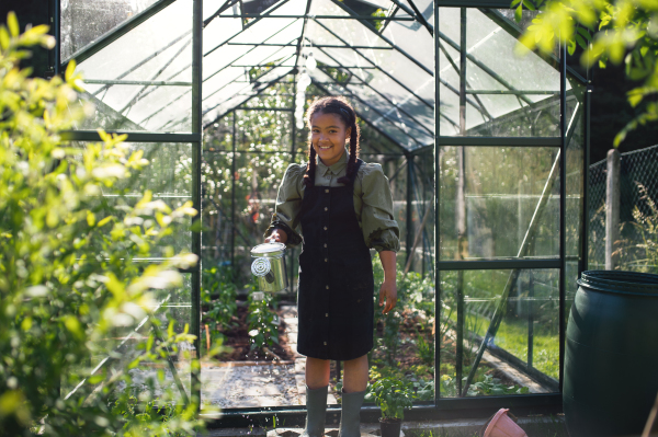 A happy small girl gardening in greenhouse outdoors in backyard, looking at camera.