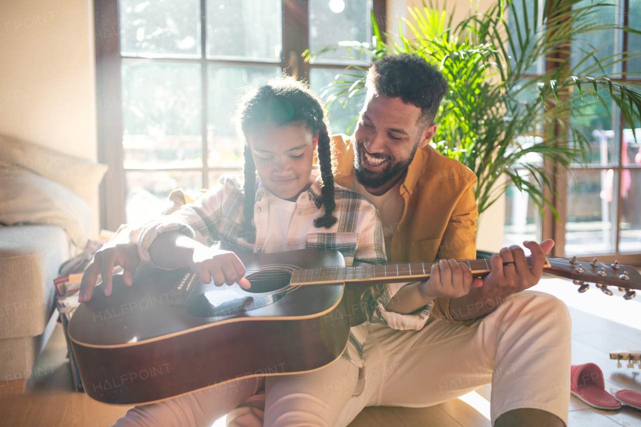 A front view of happy father with small daughter indoors at home, playing guitar.