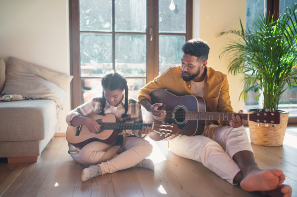 A front view of happy father with small daughter indoors at home, playing guitar.