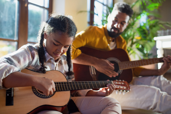 A happy father with small daughter indoors at home, playing guitar.