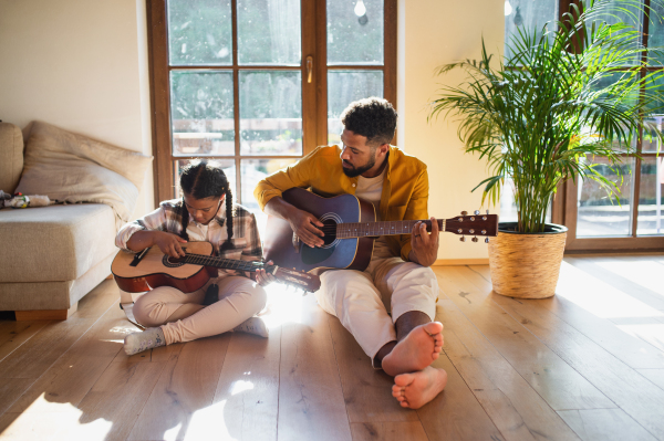 A front view of happy father with small daughter indoors at home, playing guitar.