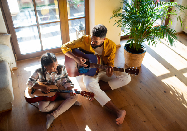 A high-angle view of happy father with small daughter indoors at home, playing guitar.