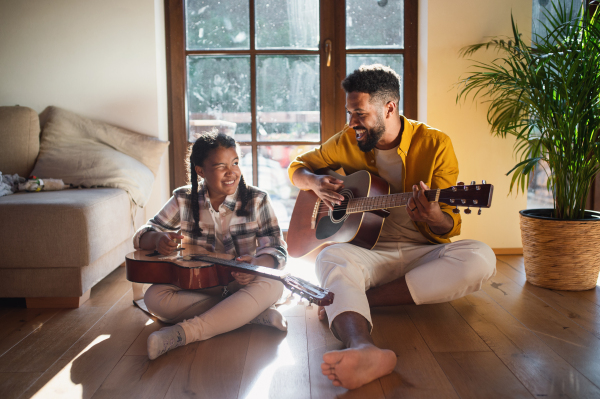 A front view of happy father with small daughter indoors at home, playing guitar.