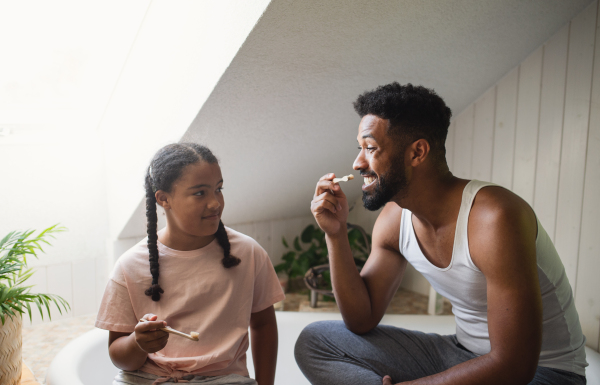 A happy young father with small daughter brushing teeth indoors at home, sustainable lifestyle.