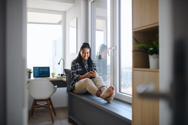 A portrait of happy teenage girl student sitting indoors in bedroom at home, using smarpthone.