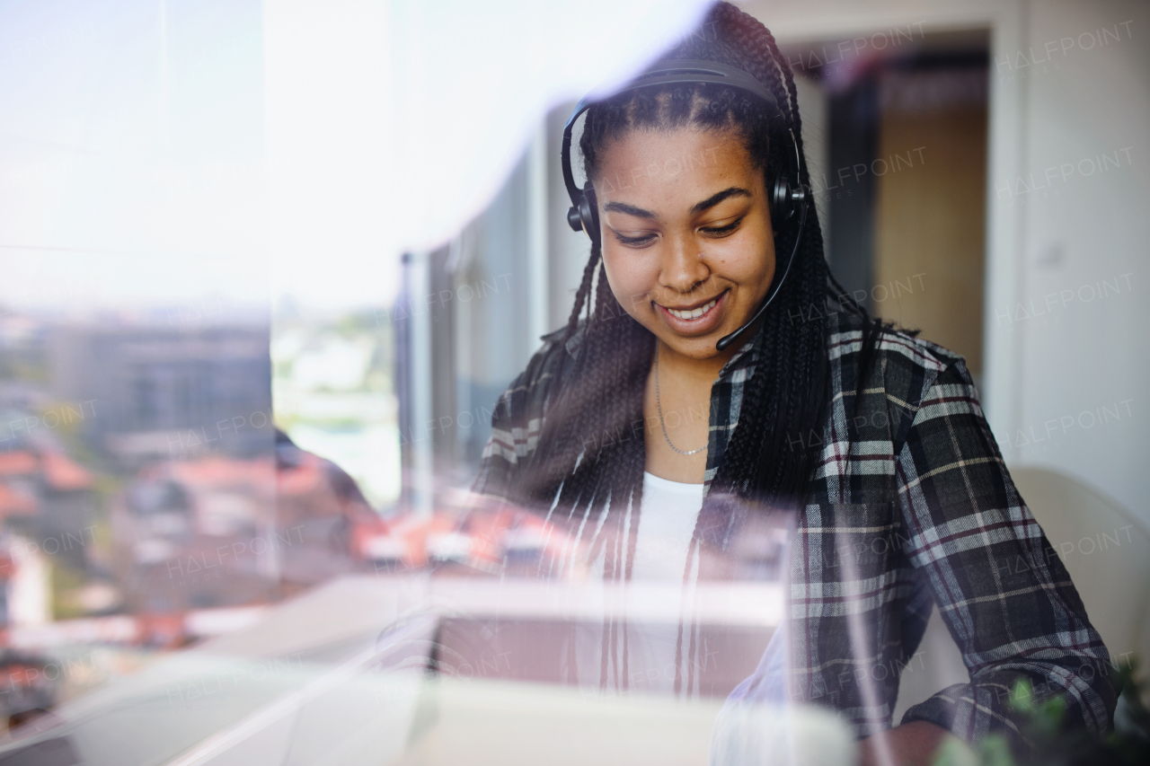 Portrait of happy teenage girl student working indoors at home, using laptop. Shot through glass.